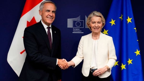 Two officials in formal attire shake hands in front of EU flags and a Canadian flag, with a European Commission backdrop.