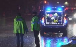 Nighttime roadside emergency: two responders in bright jackets beside an open SUV trunk with flashing blue lights on a wet road.