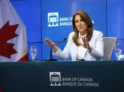 Woman in a light blazer speaks at a Bank of Canada press conference, with the Canadian flag in the background.