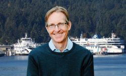Smiling man with glasses in a blue sweater stands in front of a harbor with white ferries and a forested hillside behind him.