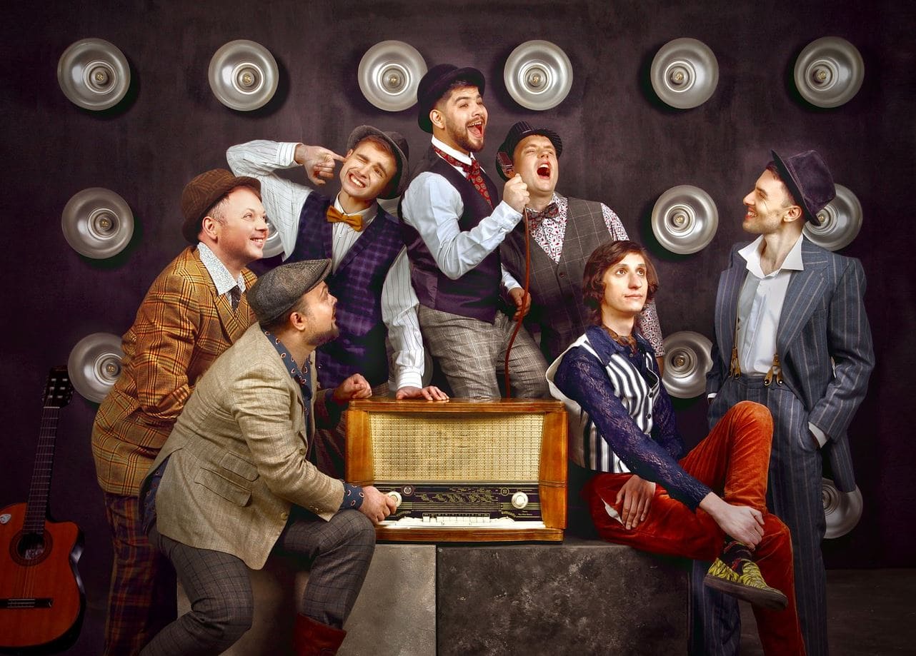 A vintage-themed band posing around an old radio, with guitars and retro outfits on a dark backdrop.