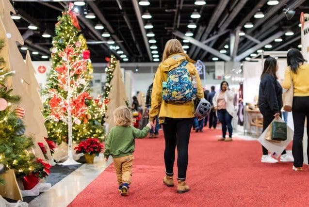 Adult and child walk hand in hand down a red-carpet aisle at a holiday market, Christmas trees with lights on the left.
