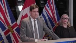 Man in a gray suit speaks at a press conference, with microphones and flags behind him; a woman with glasses sits to his right.