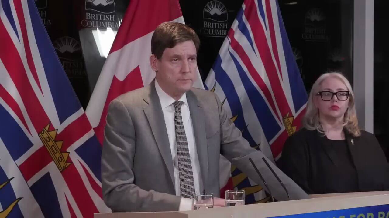 Man in a gray suit speaks at a press conference, with microphones and flags behind him; a woman with glasses sits to his right.
