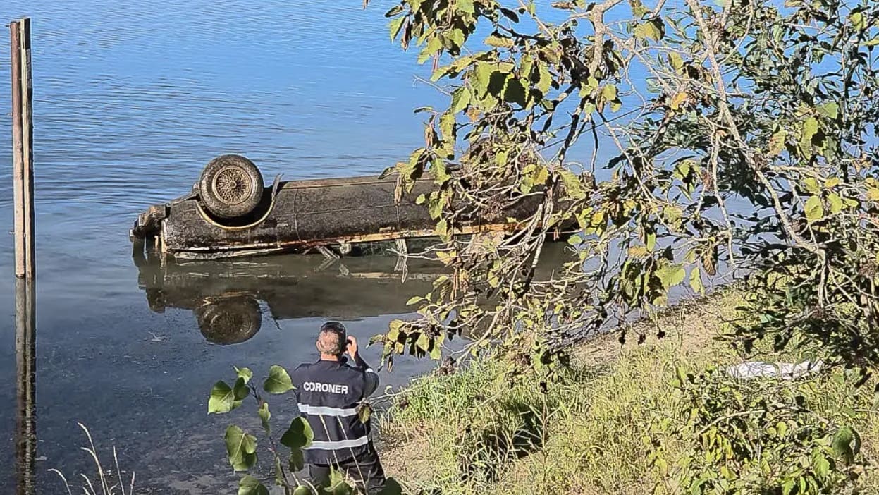 Coroner in a dark uniform standing at the water's edge, photographing an overturned boat by a calm river.