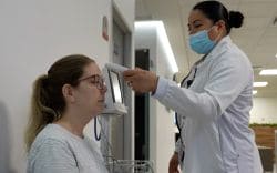 Healthcare worker in a mask takes a patient's temperature with a handheld thermometer in a clinic conference room or hallway.