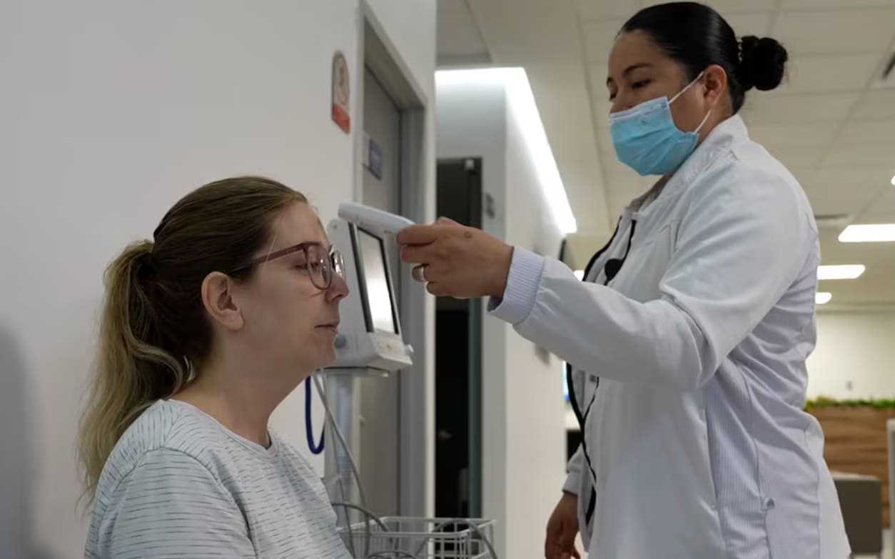 Healthcare worker in a mask takes a patient's temperature with a handheld thermometer in a clinic conference room or hallway.