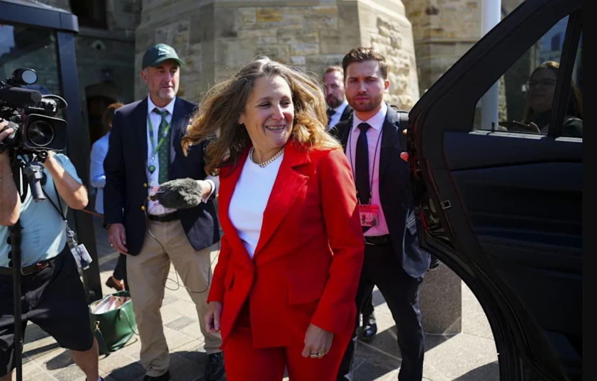 Woman in a red suit stepping from a black car as photographers film nearby outside a stone building.