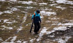 Person wearing a blue jacket, helmet, and goggles walks on a snow-dusted, muddy slope carrying skis and ski poles.