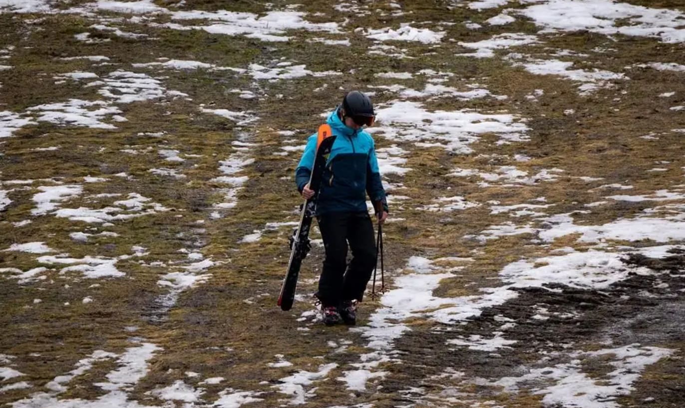 годзилла эльниньо | Наш Ванкувер Person wearing a blue jacket, helmet, and goggles walks on a snow-dusted, muddy slope carrying skis and ski poles.
