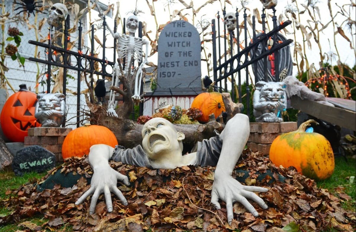 Halloween yard display with a life-size zombie crawling from a pile of leaves among pumpkins, skeletons, and a tombstone backdrop.
