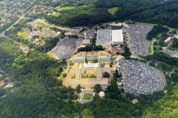 Aerial view of a university campus with terraced central buildings surrounded by dense trees and large parking lots on either side.