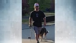 Man in a black shirt and cap walks a skateboard on a sunny street, with a dog walking beside him.