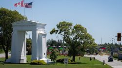 White triumphal arch gateway at a border crossing with Canadian and American flags, green toll booths, trees, and a multi-lane road nearby.