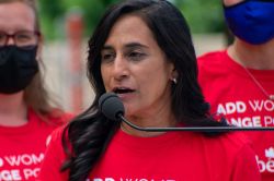 Woman in a red shirt speaks into a microphone at an outdoor rally, with masked supporters in the background on a sunny day.