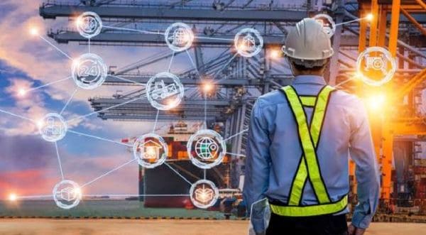 Port worker with a safety vest and hard hat stands with back to camera, facing a cargo yard with a network of glowing holographic icons around him, symbolizing connected logistics.