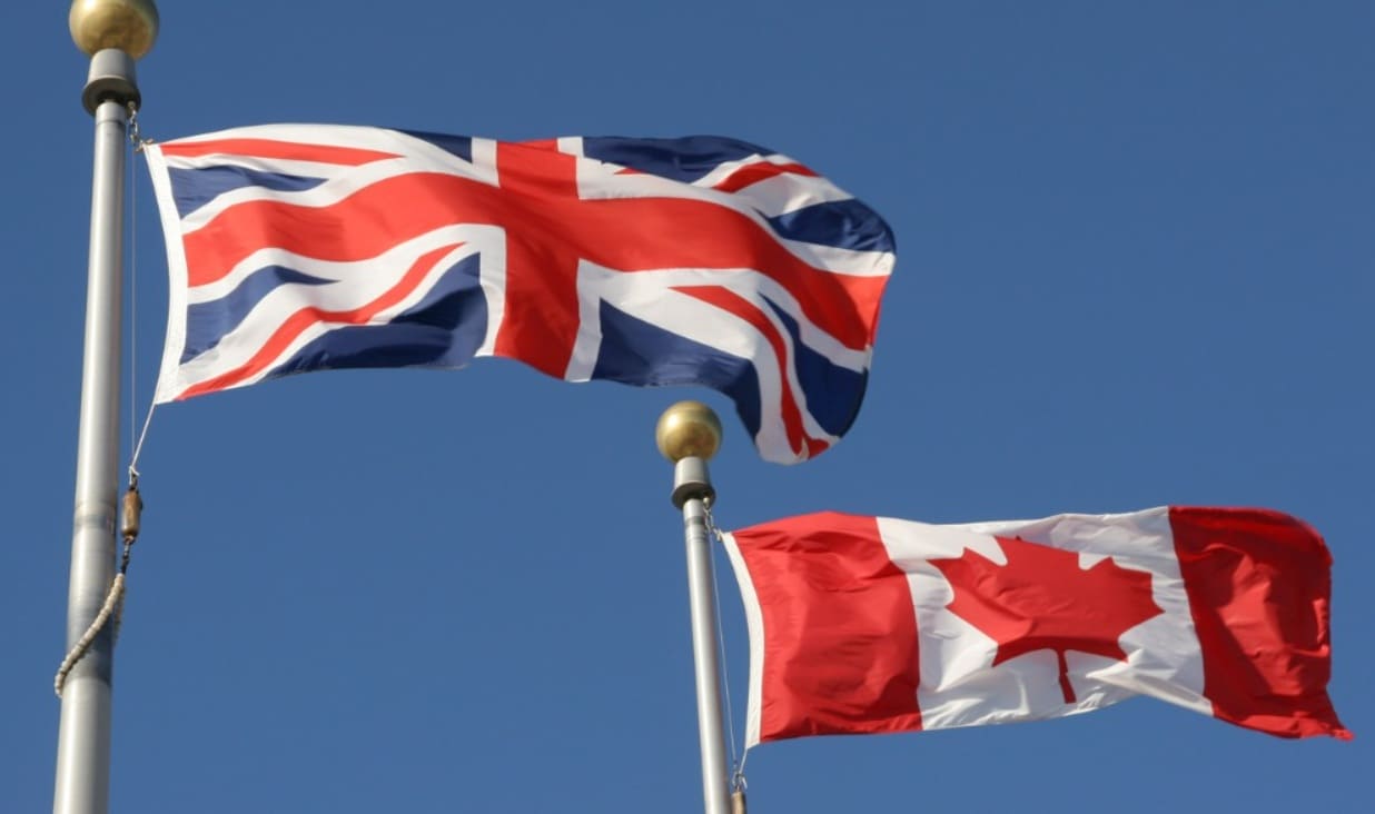 Union Jack and Canadian flag waving on flagpoles against a clear blue sky.