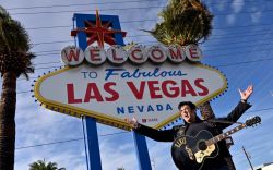 Elvis impersonator posing with a guitar beside the Welcome to Fabulous Las Vegas sign.