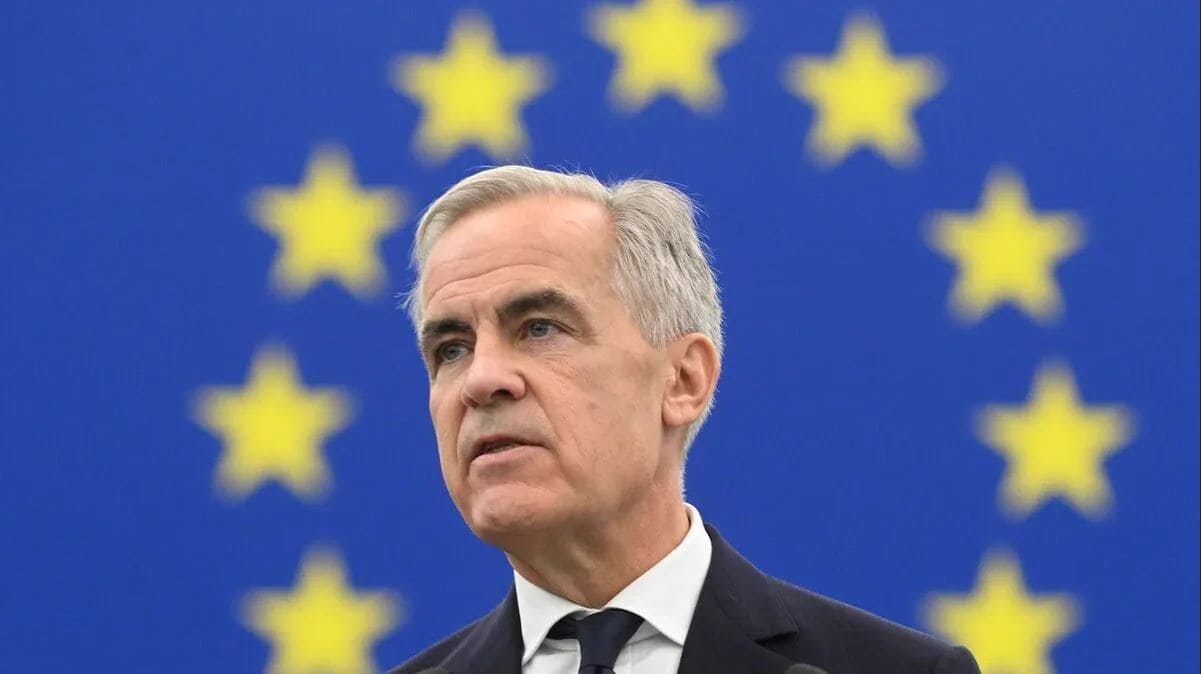Канаде грозит введение углеродного налога | Наш Ванкувер Man in a dark suit and tie stands in front of a blue European Union flag with yellow stars, looking serious.