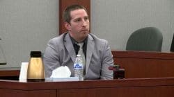 Man in a gray suit and tie sits at a courtroom desk, with a water bottle and tissue box in front.