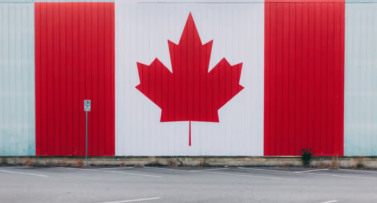 канаские технологии 2026 | Наш Ванкувер Large mural of the Canadian flag painted on a building, with red panels and a central white stripe featuring a red maple leaf, in a parking lot.