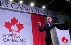 Politician in a dark suit giving a speech at a podium on a stage, with a large Canadian flag backdrop and the words 'Forever Canadian' visible.