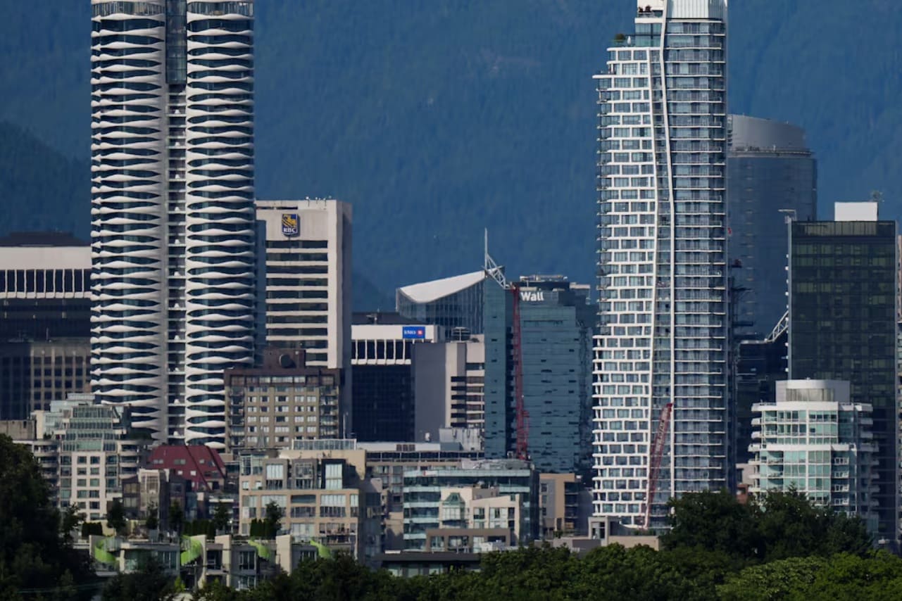 City skyline with modern high-rise buildings and a mountainous backdrop; lush green trees in the foreground.