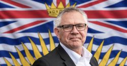 Man in a suit and glasses posing in front of a flag with a yellow sunburst and crown emblem in blue, red, and white.