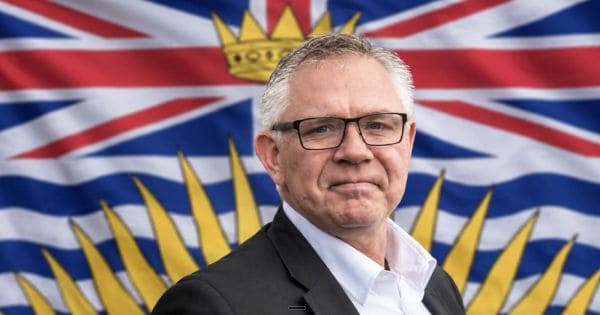 Man in a suit and glasses posing in front of a flag with a yellow sunburst and crown emblem in blue, red, and white.