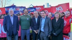 Group of diverse adults in suits and casual wear posing for a photo at a political rally, with supporters behind them holding red 'NO' signs and blue 'Enough' banners.