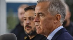 Man in a suit speaking to reporters, close-up side profile with a microphone in foreground and blurred staff in background