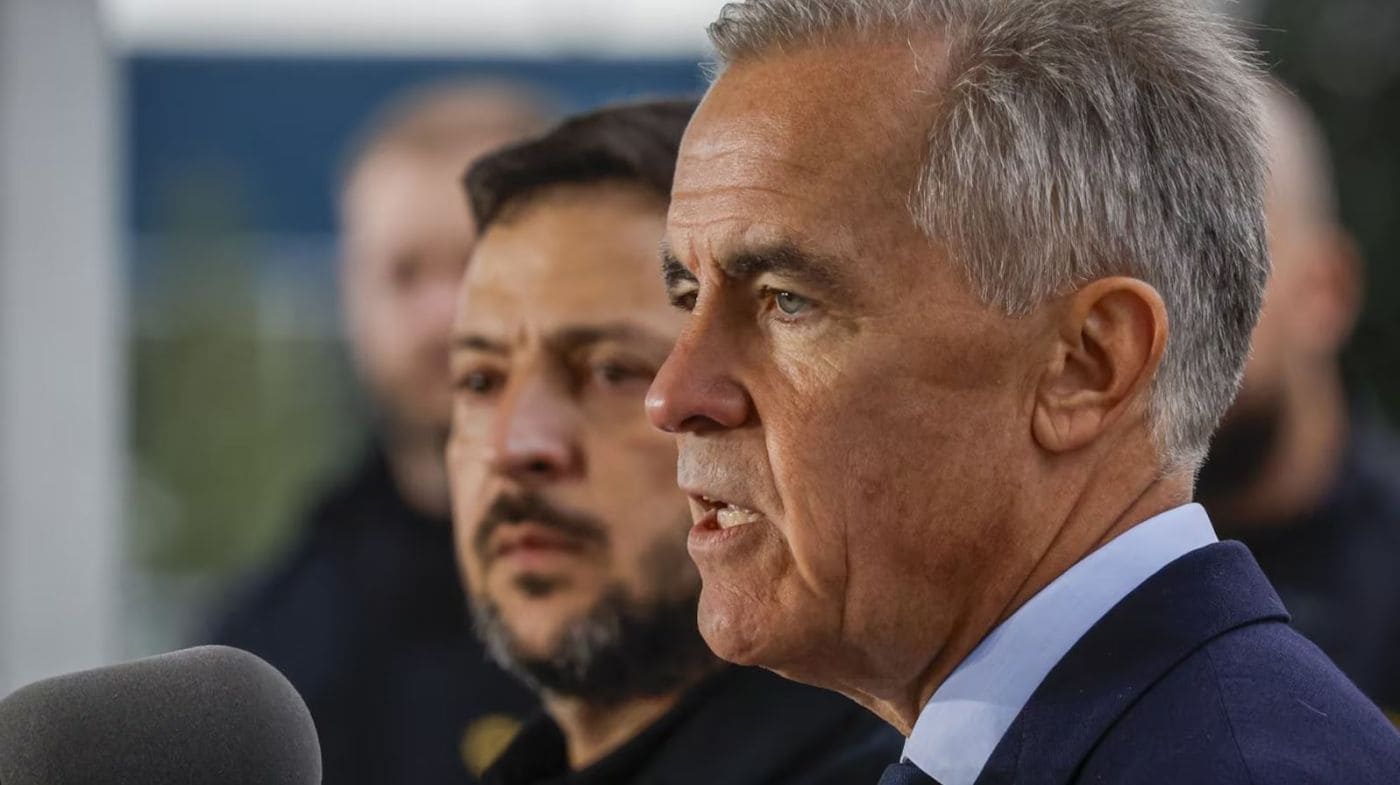 Марк Карни | Наш Ванкувер Man in a suit speaking to reporters, close-up side profile with a microphone in foreground and blurred staff in background