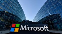 Microsoft logo on a dark sign in front of glass office buildings on a sunny day.