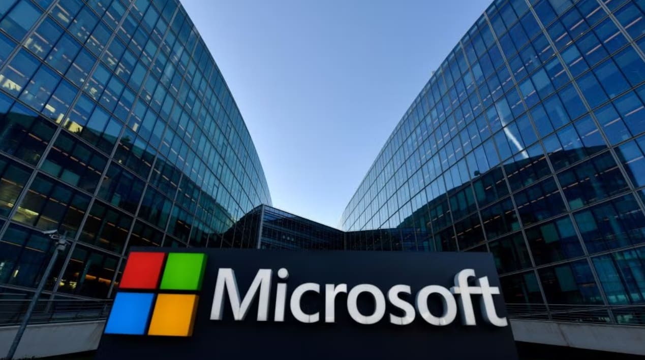 Microsoft logo on a dark sign in front of glass office buildings on a sunny day.