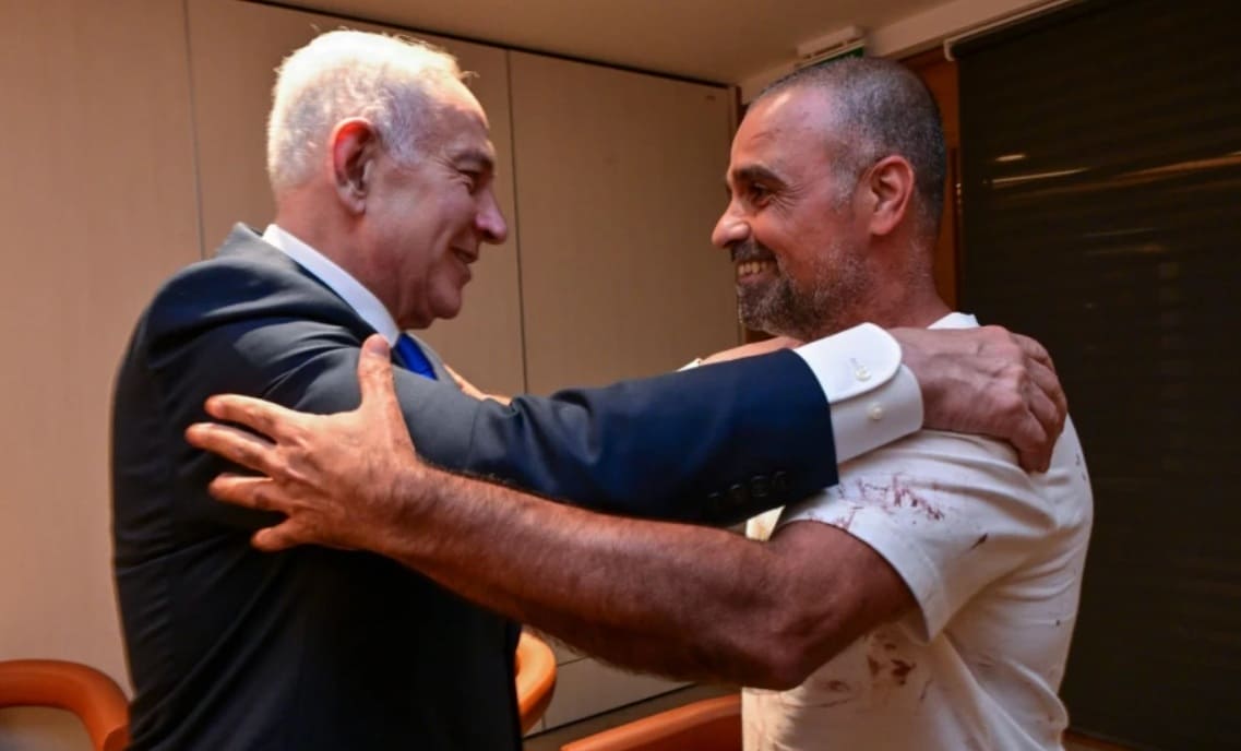Two men hug warmly in an office, smiling at each other; one in a dark suit, the other in a light shirt with red stains on the sleeve.