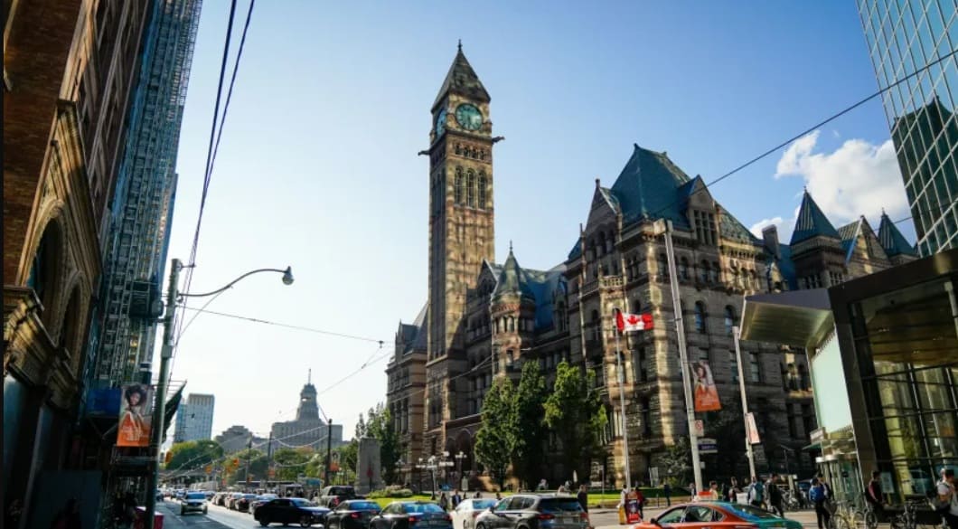 население канады стареет | Наш Ванкувер Busy city street in front of a historic building with a tall clock tower and Gothic architecture; Canadian flag displayed outside, daytime traffic nearby.