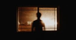 Silhouette of a person standing in front of a lit window, backlit by a warm glow behind glass panes.