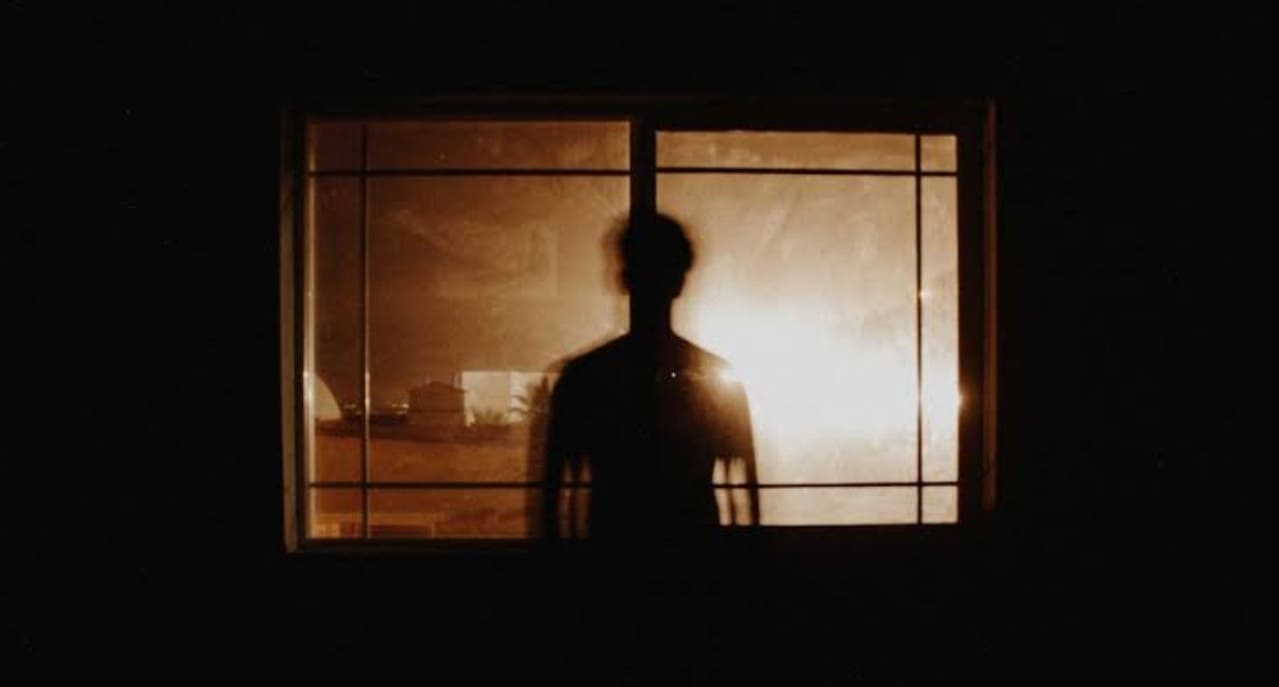 Silhouette of a person standing in front of a lit window, backlit by a warm glow behind glass panes.