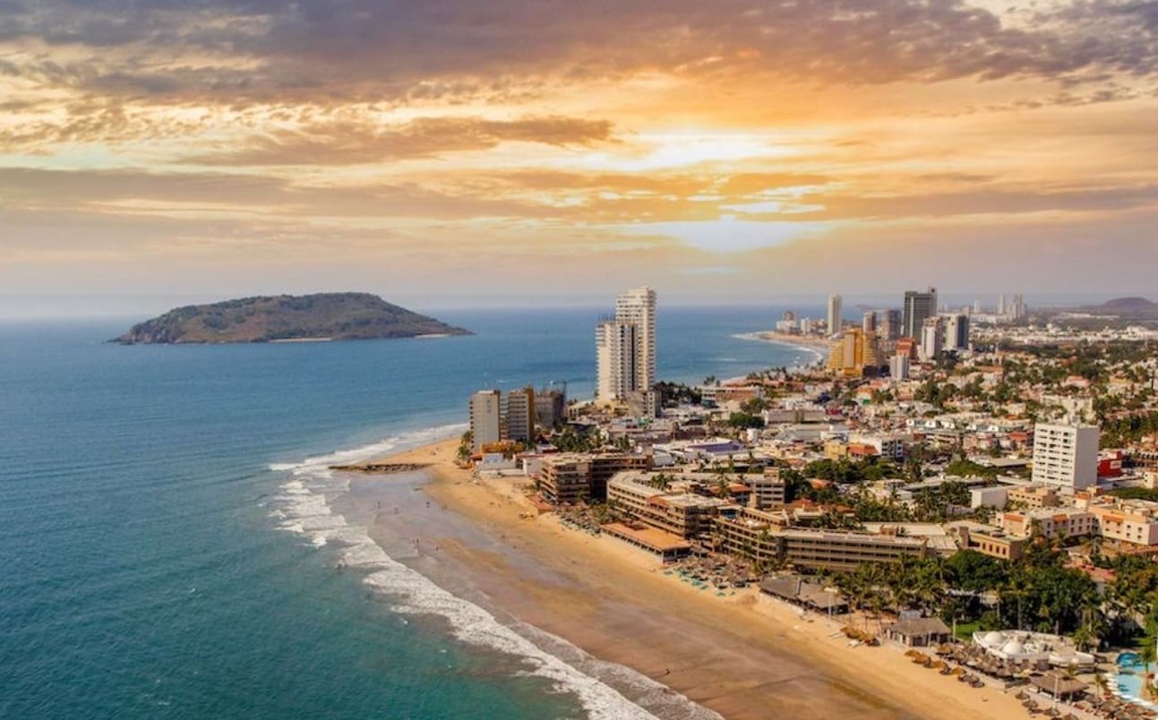 Aerial view of a coastal city at sunset with tall buildings along a sandy beach and an island in the sea to the left