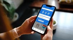 Person holding a smartphone displaying a blue shopping app interface with a search bar, delivery options, and product cards on screen.