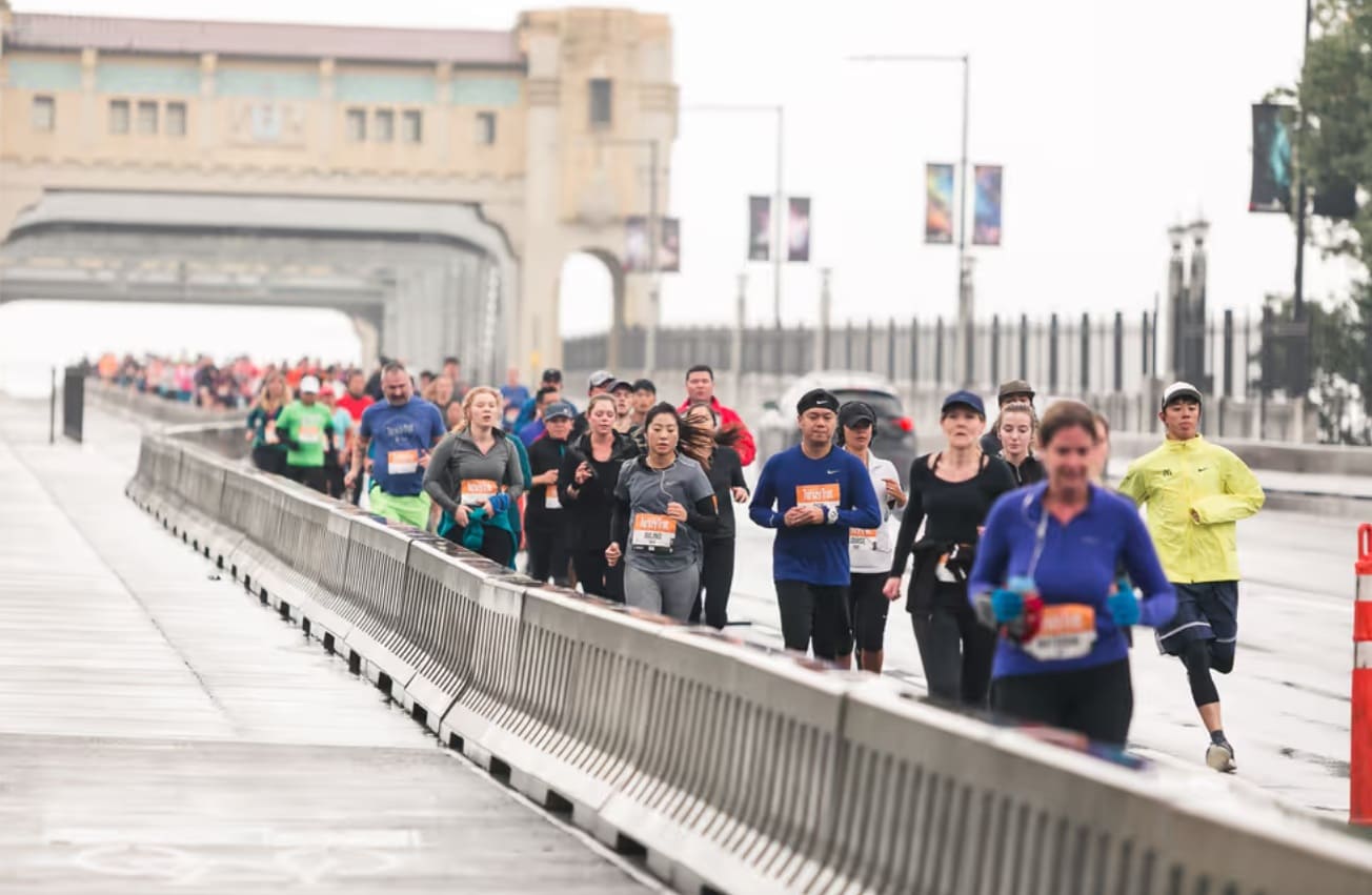 перекрытие моста | Наш Ванкувер A large group of runners in athletic gear participating in a road race along a fenced bridge on a cloudy day.