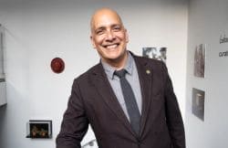 Smiling man in a dark blazer and gray shirt standing in a bright gallery space.