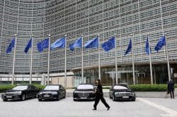 Row of European Union flags in front of a modern glass building, with black luxury cars parked along the curb and pedestrians passing by.