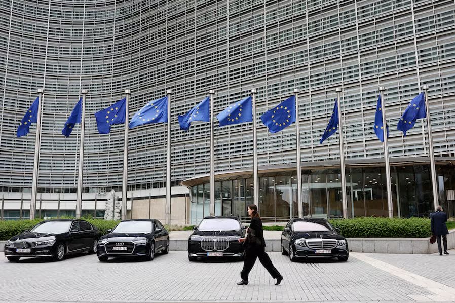 Платежные системы нового поколения | Наш Ванкувер Row of European Union flags in front of a modern glass building, with black luxury cars parked along the curb and pedestrians passing by.