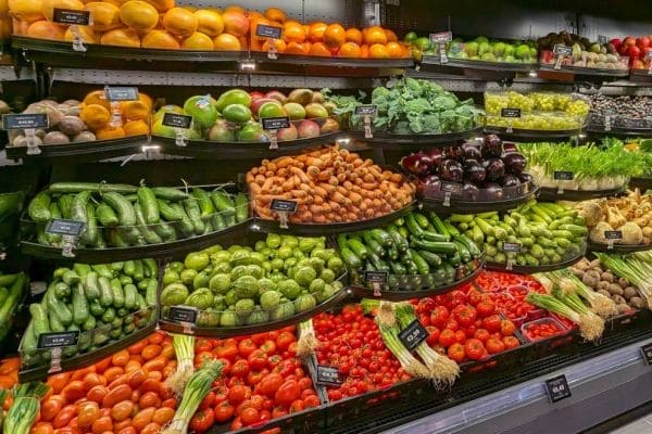 Colorful grocery store produce display with curved shelves of tomatoes, cucumbers, peppers, broccoli, and greens.