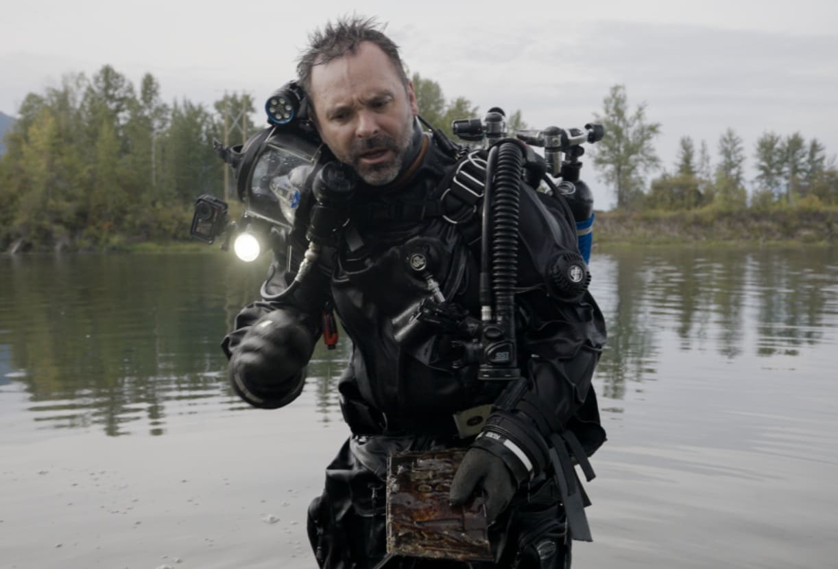 Male diver in full scuba gear emerges from a lake, holding a rusty metal artifact or box are in his hands.