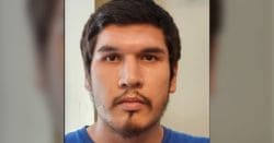 Close-up portrait of a young man with dark hair and facial hair, wearing a blue shirt, looking at the camera with a neutral expression.