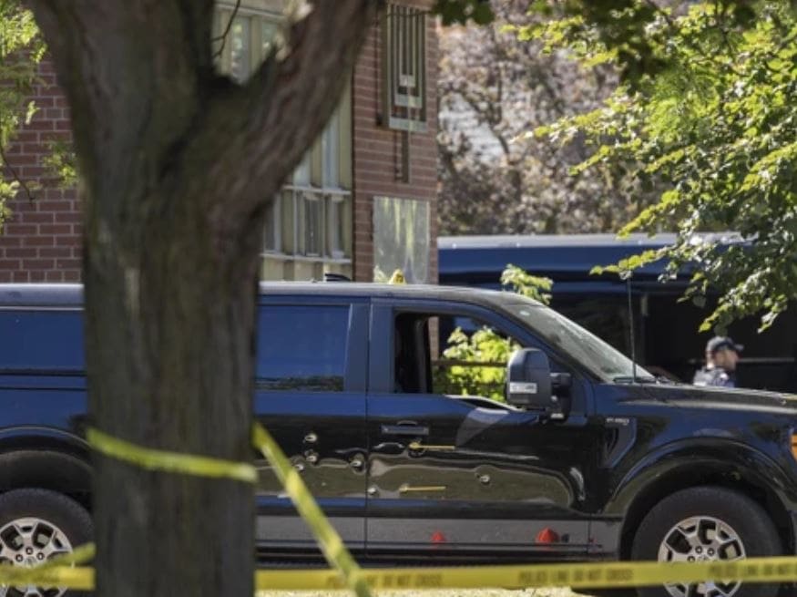 Black cargo van behind yellow caution tape in a residential area, with a tree and brick building in the background.