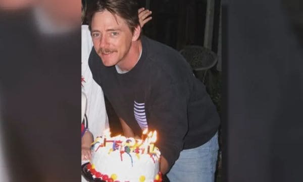 Man in a dark sweater leaning over a white-frosted birthday cake with lit candles, smiling at the camera.