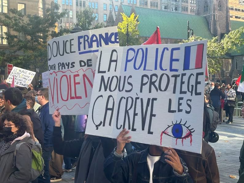 Протесты в Квебеке | Наш Ванкувер Crowd of protesters holding handmade signs in a city square; the prominent sign reads in French: 'LA POLICE NOUS PROTÈGE' with a blue eye illustration, conveying a critical message about police protection.